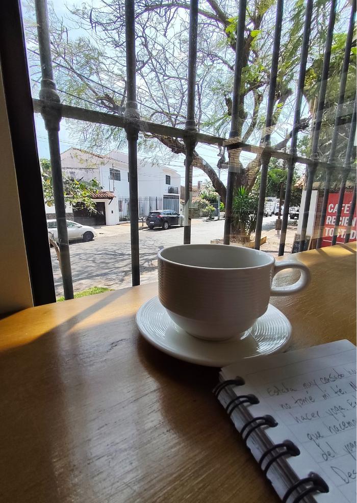 A cup of coffee on a wooden table by a window, looking out onto a quiet residential street in Paraguay.